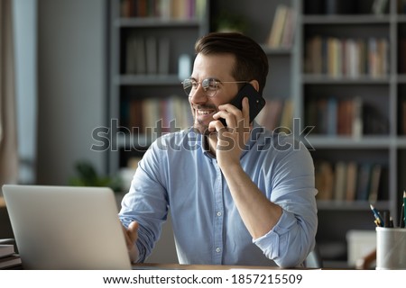 Smiling good-looking businessman in glasses sit at workplace desk in modern office room using wireless device holding smartphone talking to client, solve issues remotely. Business conversation concept Royalty-Free Stock Photo #1857215509