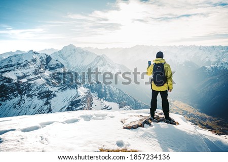 Climber take a mountain landscape photo on his smart phone. Hiker with backpack standing on top of a mountain and enjoying view. Tourist walk in the snow in the mountains during the winter holidays. 
