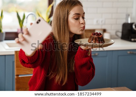 Cheerful lovely young girl taking selfie with a christmas cake while sitting at the kitchen