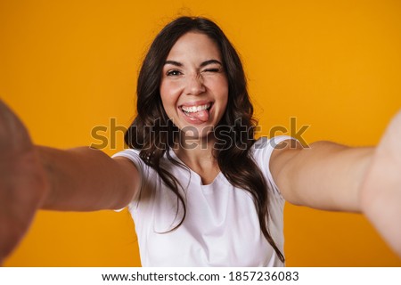 Cheerful beautiful girl showing her tongue and taking selfie isolated over yellow background