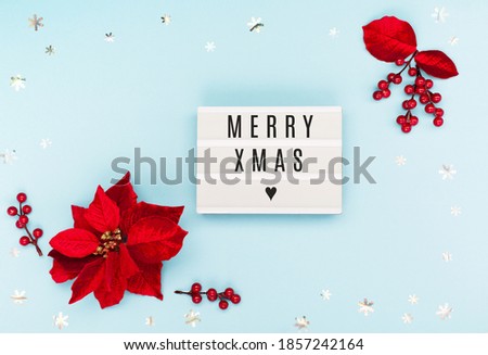 Merry Xmas text on white Lightbox with red flower poinsettia, silver snowflakes on blue paper background. Flat lay, top view, copy space.