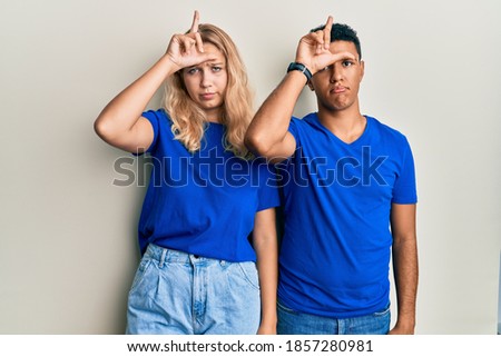 Young interracial couple wearing casual clothes making fun of people with fingers on forehead doing loser gesture mocking and insulting. 