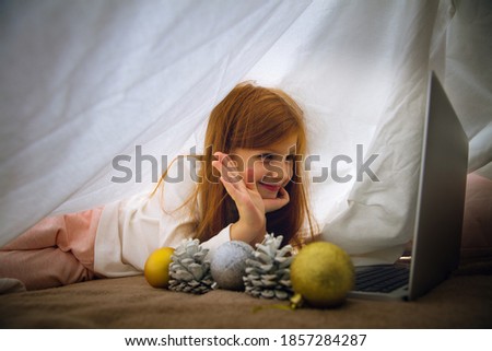 Wishes. Happy caucasian little girl during video call with laptop and home devices, looks dreamful and happy. Talking to Santa before New Year's eve, her family, watching cartoons, typing text. Bokeh