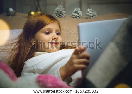 Comfortable. Happy caucasian little girl during video call with laptop and home devices, looks dreamful and happy. Talking to Santa before New Year's eve, her family, watching cartoons, typing text