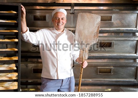 Middle-aged baker standing in his bakery with a wooden tray in his hand and smiling at the camera Royalty-Free Stock Photo #1857288955