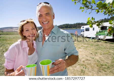 Horizontal shot of a mature couple with mugs by motor home and lake in a park.