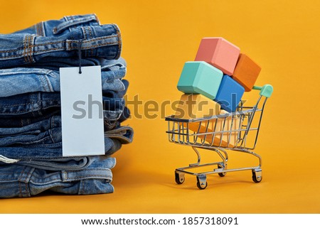 A stack of blue jeans with a white blank tag on a yellow background. Shopping trolley with multi-colored cubes. Sales consept