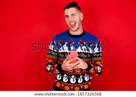 Young handsome Caucasian man wearing Christmas sweater against red wall,  taking a selfie  celebrating success