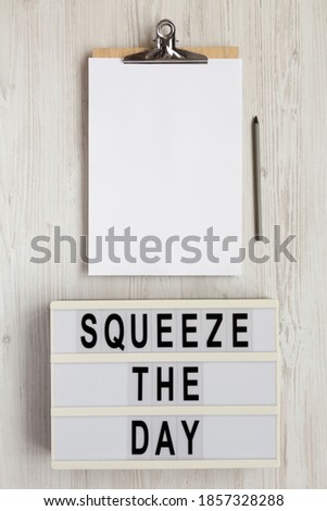 'Squeeze the day' on a lightbox, clipboard with blank sheet of paper on a white wooden background, overhead view. Flat lay, top view, from above. 