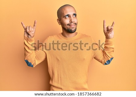 Hispanic adult man doing rock gesture over yellow background smiling looking to the side and staring away thinking. 