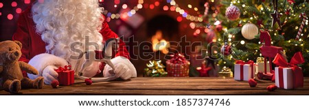 Santa Claus Sits At A Table With Gifts And Toys Near The Fireplace And Christmas Tree