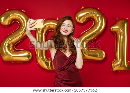 Smiling stunning young redhead woman in elegant dress doing selfie shot on mobile phone isolated on red background, golden numbers air balloons. Happy New Year 2021 celebration holiday party concept