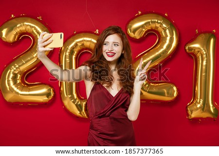 Smiling young woman in elegant dress doing selfie shot on mobile phone showing victory sign isolated on red background, golden numbers air balloons. Happy New Year 2021 celebration holiday concept
