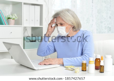 Portrait of ill beautiful senior woman with facial mask using laptop