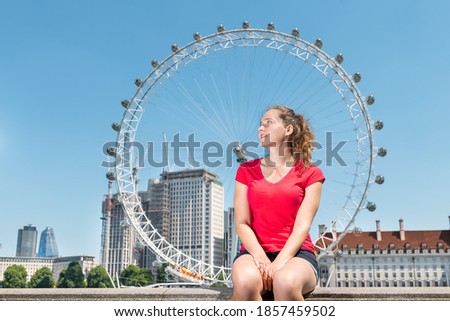 Young happy woman sitting on railing looking at cityscape skyline of city with Thames River and London Eye in United Kingdom during sunny summer