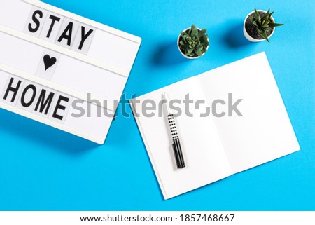 White lightbox with text STAY HOME and notebook on blue background. Online learning, working, social distancing concept