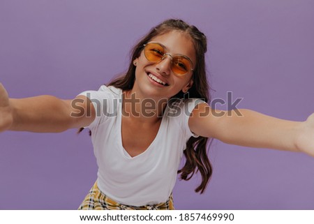 Happy tanned young girl takes selfie on purple background. Attractive teenager in white t-shirt smiles on isolated.