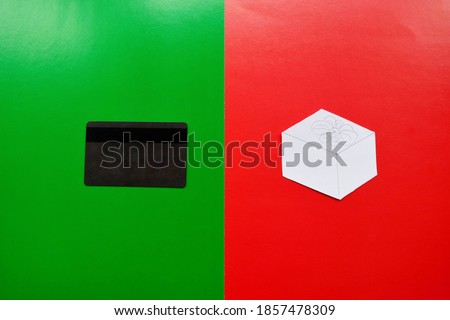 A credit card on the green half and a paper figurine of a gift box on a red background, opposite each other. Conceptual photo of shopping for gifts online.
