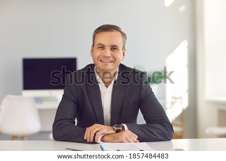 Portrait of young smiling businessman in suit sitting and looking at camera during online videocall in office over modern company interior at background. Working in office, business, online concept Royalty-Free Stock Photo #1857484483