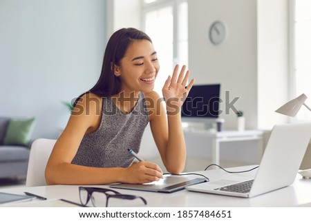 Smiling creative young digital artist girl with graphic tablet and pen waving hand at laptop computer, meeting client online from home office and painting or drawing sketch portrait via video call app Royalty-Free Stock Photo #1857484651