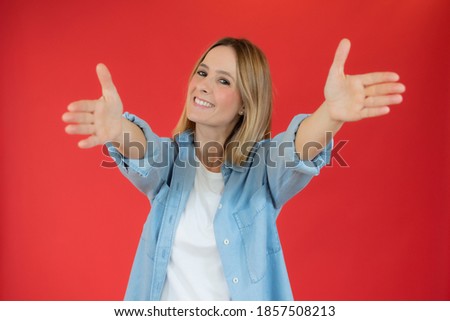 Young beautiful woman wearing denim shirt over red background looking at the camera smiling with open arms for hug. Cheerful expression embracing happiness.
