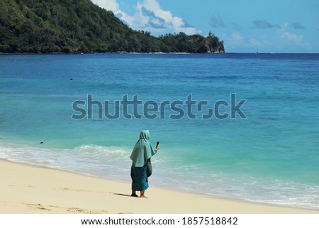A woman stand by the beach while taking picture with her smartphone