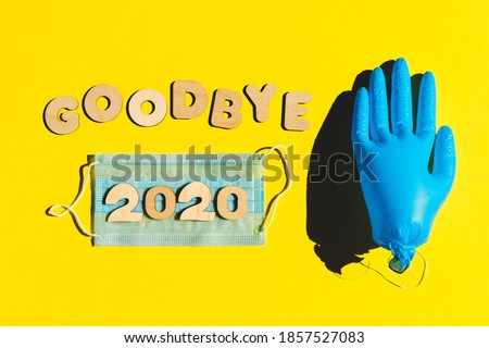 Words GOODBYE 2020 from wooden letters, inflated medical glove waving bye-bye and facemask on a yellow background. Year 2020 and epidemic concept. Royalty-Free Stock Photo #1857527083