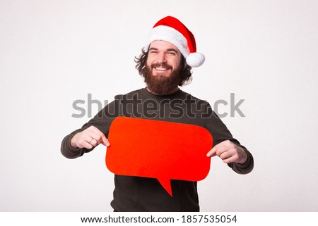 Charming young man with beard smiling and holding red speech bubble, Wearing santa claus hat