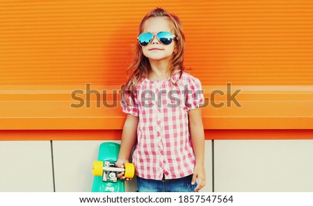 Portrait of stylish little girl child with skateboard in the city over an orange background