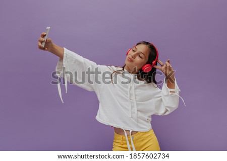 Stylish teenager listens to music in headphones on purple background. Happy young girl takes selfie and shows peace sign.