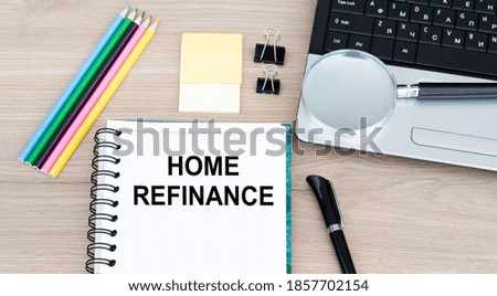 Text HOME REFINANCE on an open notebook on an office table, next to a laptop, a pen, a magnifying glass.
