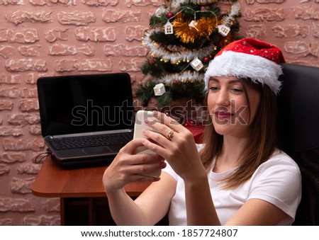 The girl communicates via smartphone video link. New year's mood, cap on the head, Christmas tree with decorations. Selective focus.