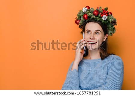Beautiful smiling woman wears Christmas wreath, talking on mobile phone conducting pleasant conversation, isolated on orange studio wall with copy space for your advertising content. Happy New Year