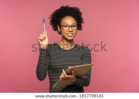 Young happy Afro-American teacher woman isolated on pink studio wall. Student girl wear glasses holding folder and pen, smiling, looking at camera. Education in high school university college concept