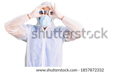 Young hispanic man wearing doctor protection coronavirus uniform and medical mask doing ok gesture like binoculars sticking tongue out, eyes looking through fingers. crazy expression. 