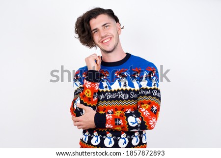 Cheerful Young handsome caucasian man wearing Christmas sweater against white background, with hand near face. Looking with glad expression at the camera after listening to good news. Confidence.