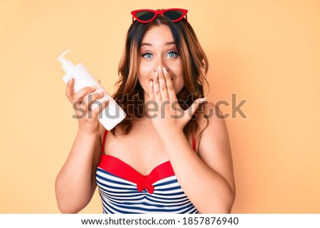 Young beautiful caucasian woman wearing swimsuit and holding sunscreen lotion covering mouth with hand, shocked and afraid for mistake. surprised expression 