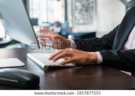 Office manager male hand typing on computer keyboard, right hand holding a glass of water, closeup. Businessman working, typing on the wireless keyboard, no face, concept of office work