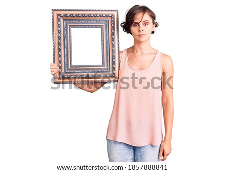 Beautiful young woman with short hair holding empty frame thinking attitude and sober expression looking self confident 