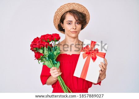 Young hispanic woman holding gift and bouquet of flowers for anniversary relaxed with serious expression on face. simple and natural looking at the camera. 