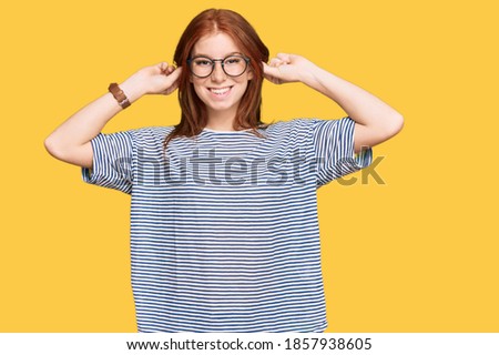Young read head woman wearing casual clothes and glasses smiling pulling ears with fingers, funny gesture. audition problem 
