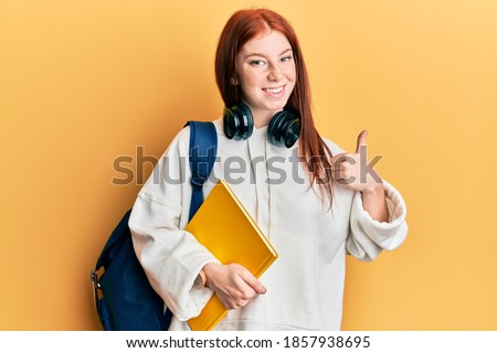 Young red head girl wearing student backpack and holding book smiling happy and positive, thumb up doing excellent and approval sign 