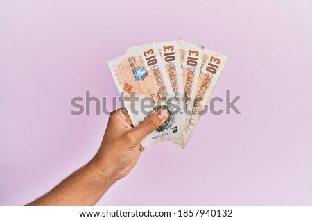 Hispanic hand holding 10 uk pounds banknotes over isolated pink background. Royalty-Free Stock Photo #1857940132