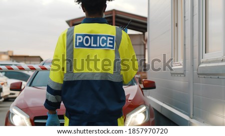 Back view of police officer in uniform with walkie talkie near car checkpoint. Driver in car giving documents to staff in booth with barrier. Toll booth with car.