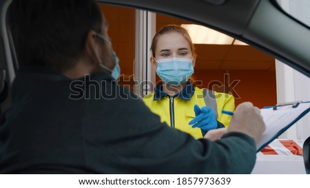 Man driver in mask signing document on clipboard entering private area. Woman worker wearing protective mask in parking lot booth giving driver paper to sign before entering