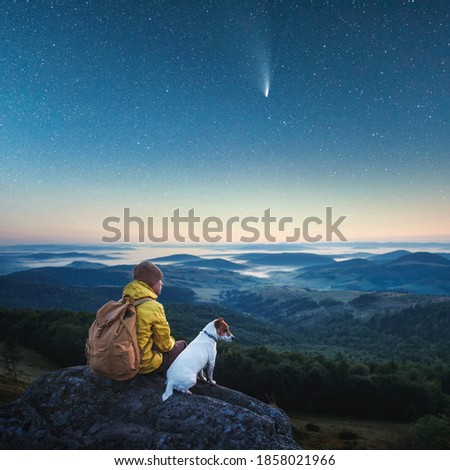 Alone tourist sitting on the edge of the cliff with white dog against the backdrop of an incredible mountains with starry night sky. Landscape photography