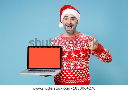Cheerful Northern bearded man frozen face in Santa hat Christmas sweater hold laptop computer with blank empty screen showing thumb up isolated on blue background. Happy New Year winter time concept