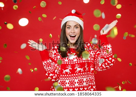 Surprised young Santa woman 20s wearing sweater Christmas hat celebrating with confetti spreading hands isolated on red background studio portrait. Happy New Year celebration merry holiday concept