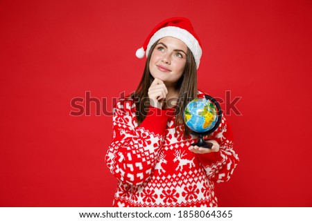 Pensive young Santa woman wearing sweater Christmas hat hold Earth world globe put hand prop up on chin isolated on red background studio portrait. Happy New Year celebration merry holiday concept