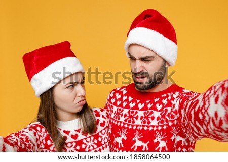 Close up offended young Santa couple friends man woman in red sweater, Christmas hat doing selfie shot on mobile phone isolated on yellow background. Happy New Year celebration merry holiday concept